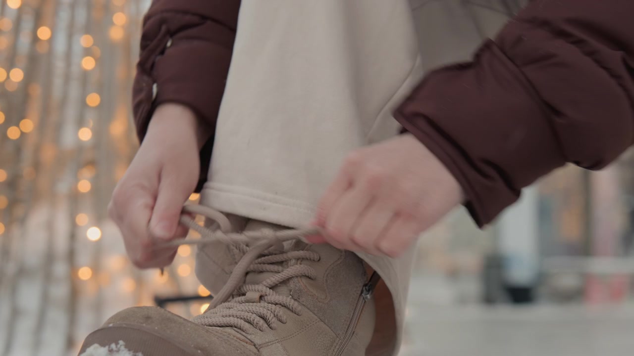Primo piano mostra mani che allacciano e stringono i lacci su uno stivaletto marrone testurizzato mentre si è seduti, con le dita che fanno passare il laccio e stringono il nodo; pantaloni beige con risvolto, manica scura e luci urbane soffuse.