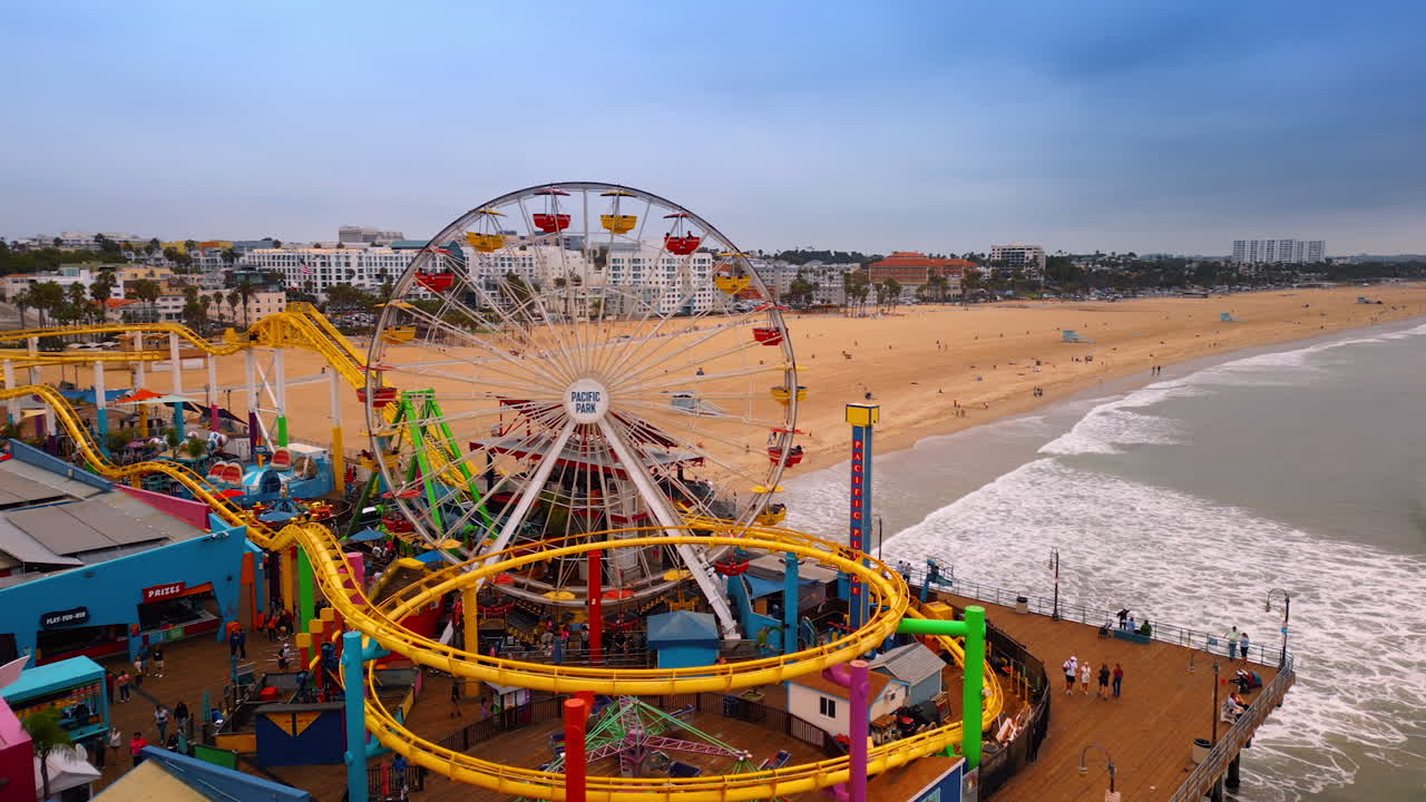 Los Angeles, USA, 29 August 2025: Flight around the roller-coasters and Ferris Wheel at Pacific Park in Santa Monica, LA, California, USA. Foamy waves of the Pacific Ocean roll to the sandy beach at backdrop