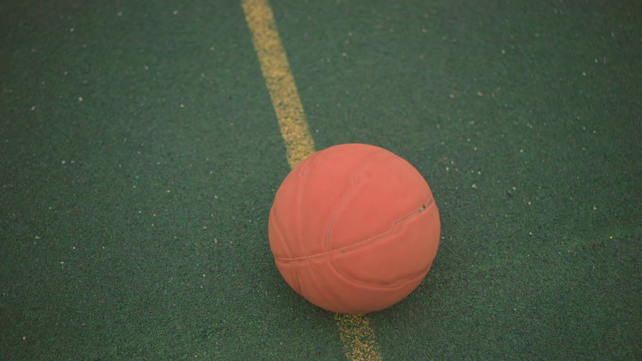 Oransje basketball på grønn bane, slitt gummiball som hviler på falmet gul linje, tom utendørs lekeplass under overskyet himmel, kornete tekstur, dempede toner, minimal filmatisk komposisjon som fanger