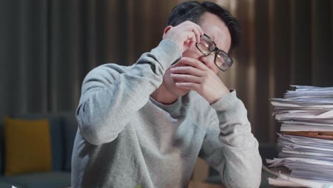 close up of tired asian man yawning and taking off his glasses before sleeping while working hard with documents at home