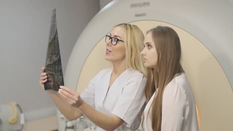 woman doctor radiologist explains the results of mri scanning for young female patient, showing the snapshot with images, observing and analyzing mri scan in modern clinic beside modern mri scanner