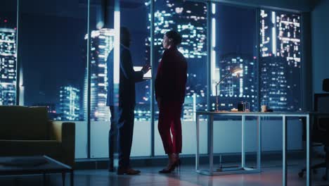 successful corporate top ceo's talking, using laptop computer standing in big city office at night. two successful managers discuss environmental, social side of e-commerce corporation