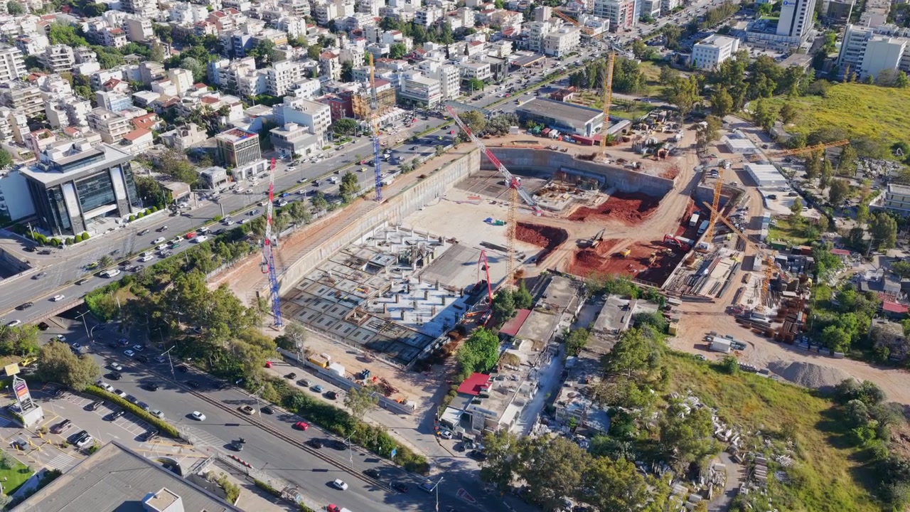 Imágenes de dron en picado mostrando una enorme obra de construcción en una ciudad bulliciosa, con grúas y maquinaria pesada trabajando en los cimientos de un nuevo edificio rodeado de tráfico denso.