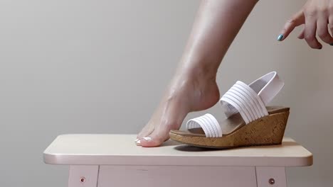 woman places feet angling into white elastic band of cork heels on stool