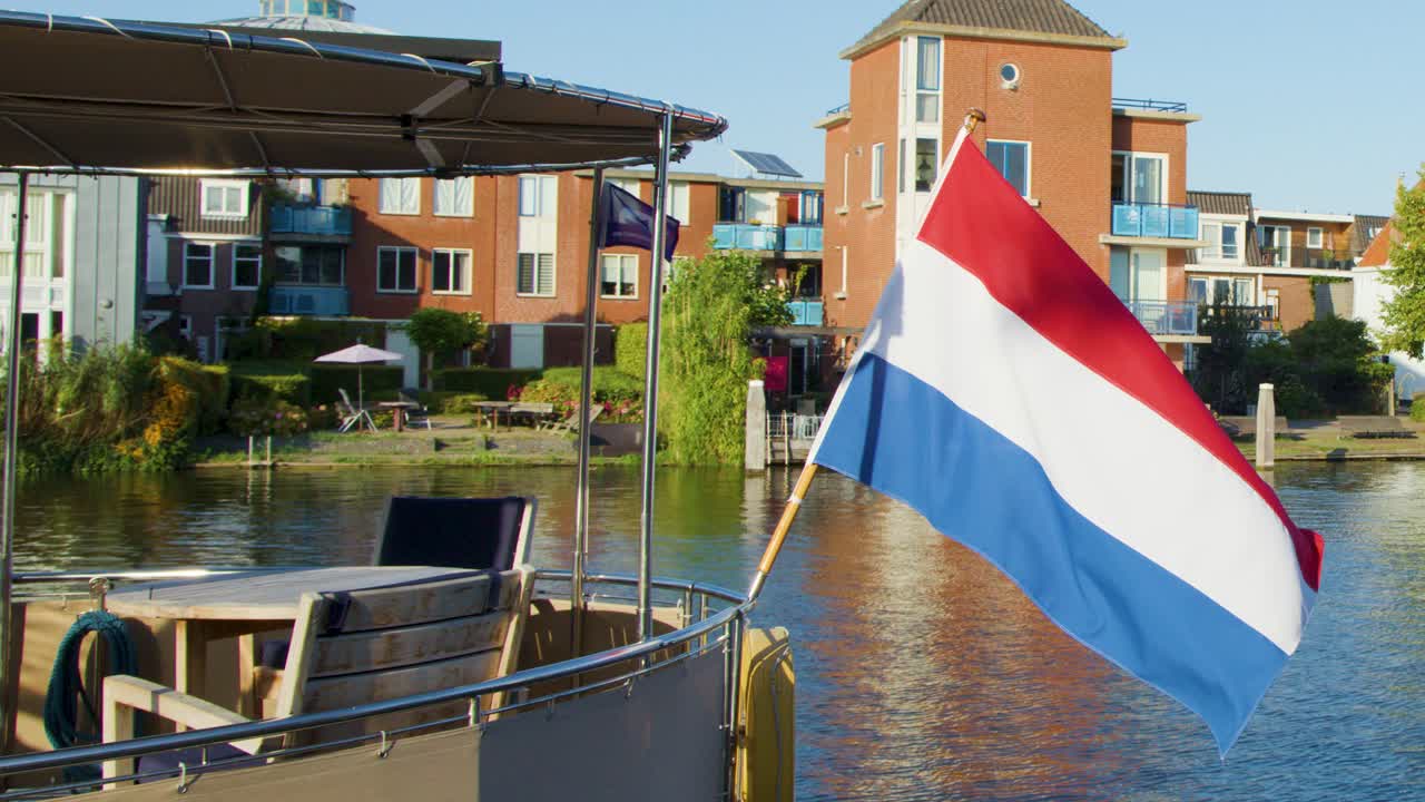 Dutch flag waves on boat along Haarlem canal, historic brick buildings, calm water, sunny day