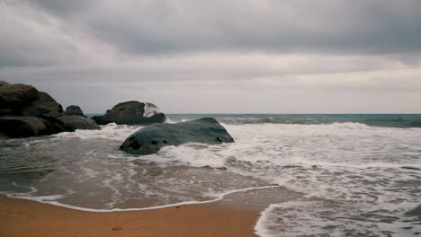 Wasser,-Das-An-Einem-Bewölkten-Tag-Mit-Blauem-Meer-Auf-Den-Felsen-An-Einer-Küste-Spritzt