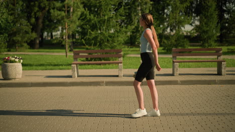 side view of young lady walking along interlocked path while holding roller skates in her hand, sunlight casts her shadow on the ground, with background featuring trees, greenery and benches