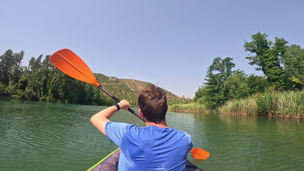A wet man is seen kayaking through a serene water body in Catalonia, Spain, surrounded by lush green trees and distant hills under a clear blue sky