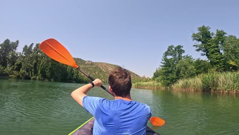 a wet man is seen kayaking through a serene water body in catalonia, spain, surrounded by lush green trees and distant hills under a clear blue sky