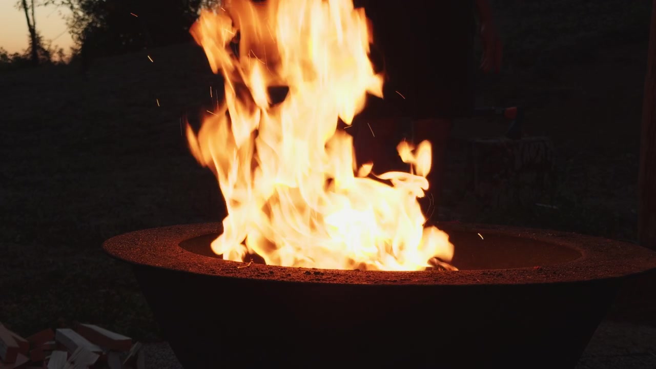 Tall orange flames rise from burning wood in a round outdoor fire pit. A stack of logs and grass surround the metal bowl in fading daylight.