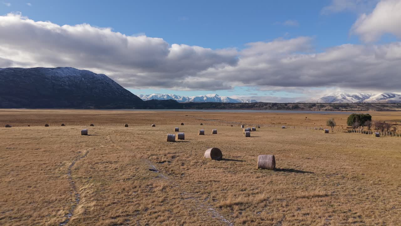 Drone pulls back over golden hay bales scattered across the Patagonian steppe—revealing the vast field beneath snow-capped Andes peaks in Chubut, Argentina, revealing farming and livestock tradition