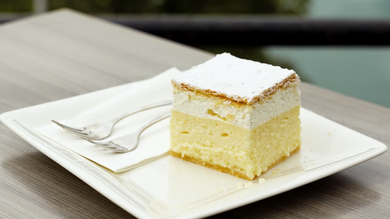 Zoom-out shot of Bled Cream Cake with white plate, fork and Lake Bled in the back in Bled, Slovenia