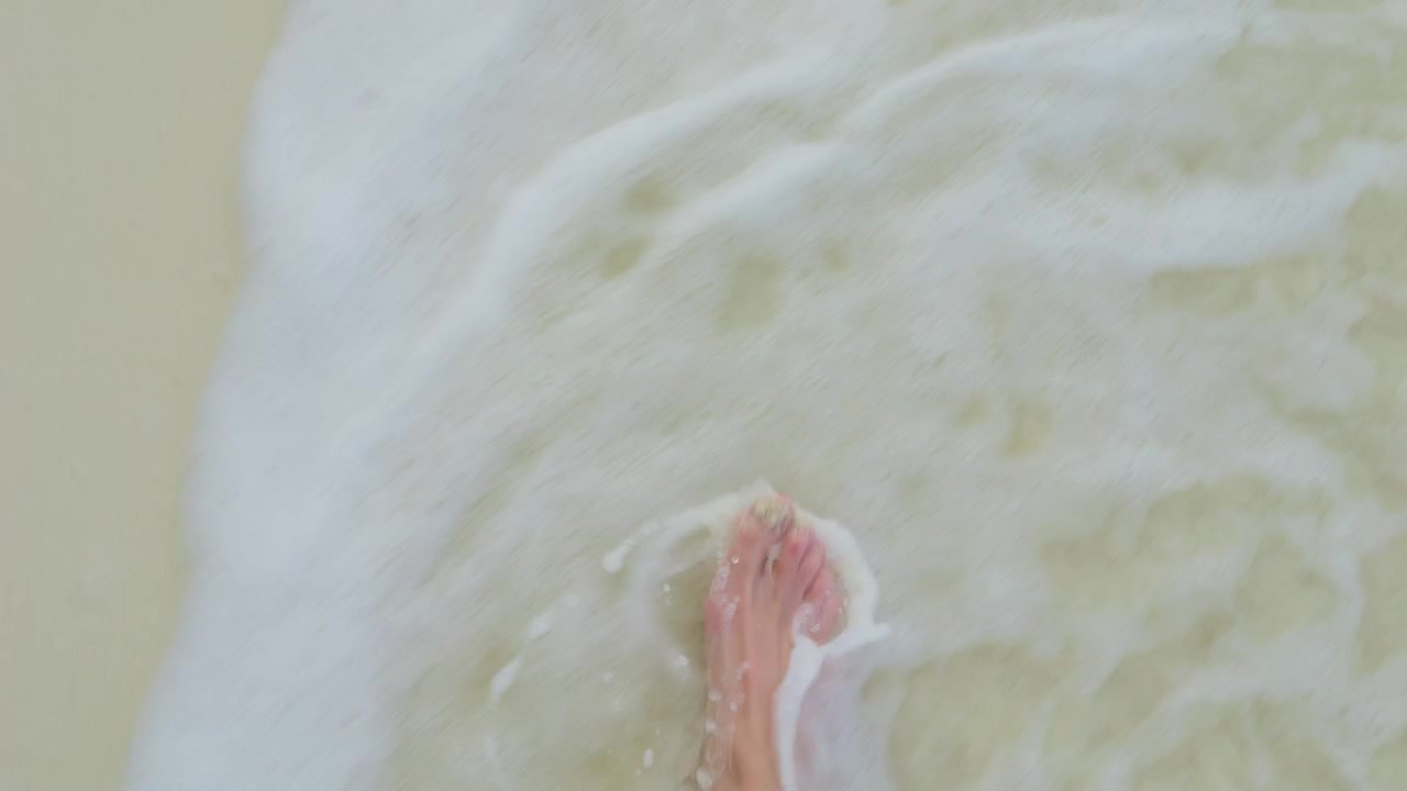Vista in prima persona di piedi che guadano un fondale sabbioso in acque poco profonde di una laguna turchese mentre una leggera risacca si infrange sulla sabbia nelle Maldive tropicali
