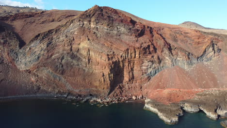 Moving-away-from-an-orange-structured-mountain-in-a-sunny-bay-Cala-Tacoron,-El-Hierro-Island