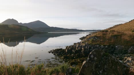 Die-Kamera-Erhebt-Sich-Sanft-über-Die-Vegetation,-Um-Die-Sonne-Zu-Zeigen,-Die-Von-Der-Oberfläche-Eines-Gezeitenseelochs-Im-Nordwestlichen-Hochland-Von-Schottland-Reflektiert-Wird,-Kurz-Bevor-Sie-Hinter-Bergen-Untergeht