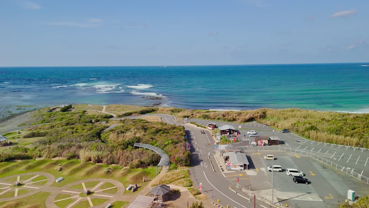 A beautiful aerial shot pans past a tall, white lighthouse to reveal the stunning turquoise ocean, a park, and a parking lot on the Japanese coast on a sunny day