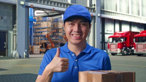 close up of asian male courier in blue uniform showing thumbs up gesture and smiling while delivering a carton, outside of logistics distributions warehouse