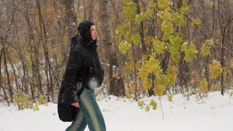 young girl in winter jacket with hands in pockets and guitar on back walking on snow-covered route in snowfall, surrounded by bare trees and falling snow