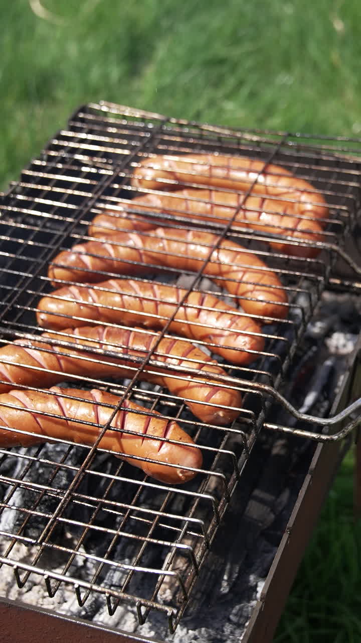 Grilling food for picnic. Delicious hot dog sausages frying in a grill grate in smoke on green nature background. Barbeque in summer. Vertical video
