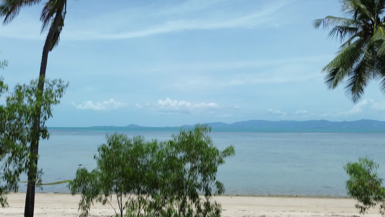 Scenic aerial drone footage of a tropical beach on Koh Phangan island, Thailand, showing palm trees, white sand, and turquoise ocean water under a clear blue sky