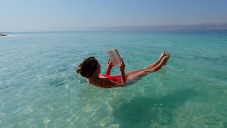 peaceful young woman floating in the dead sea wearing red swimsuit and reading a book