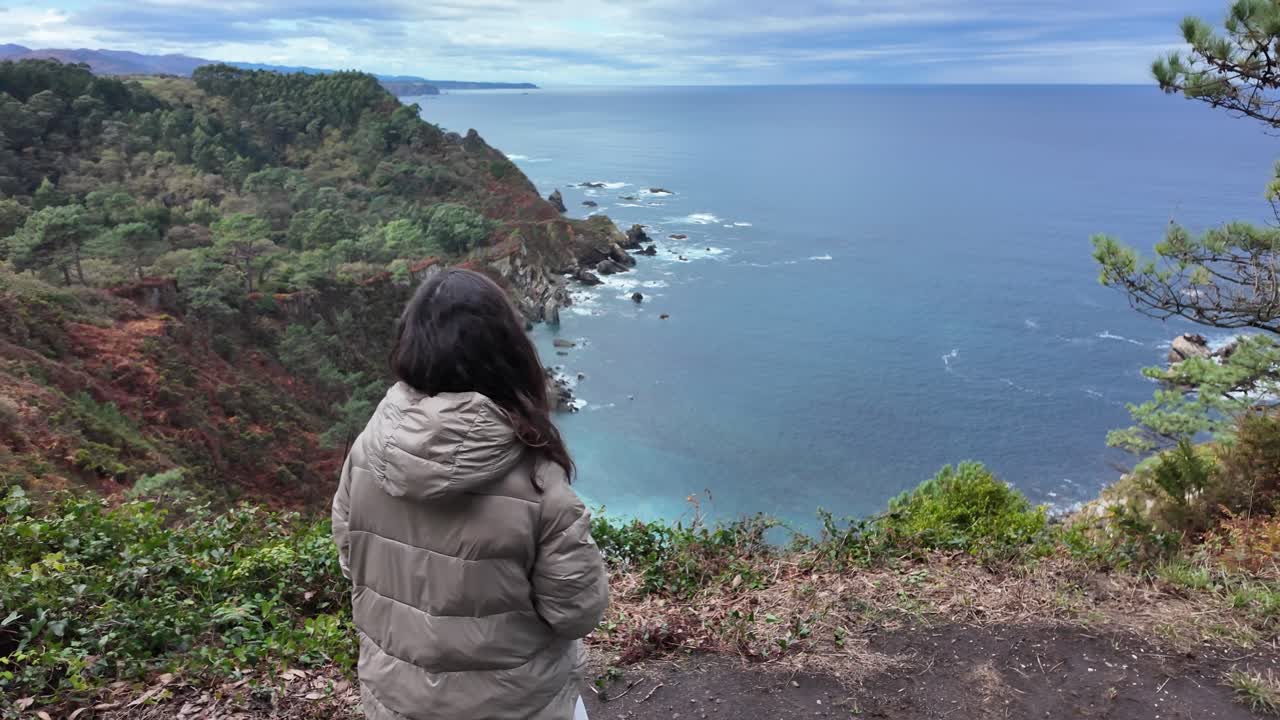 A person stands on a cliffside viewpoint, looking at the wide ocean and dramatic landscape