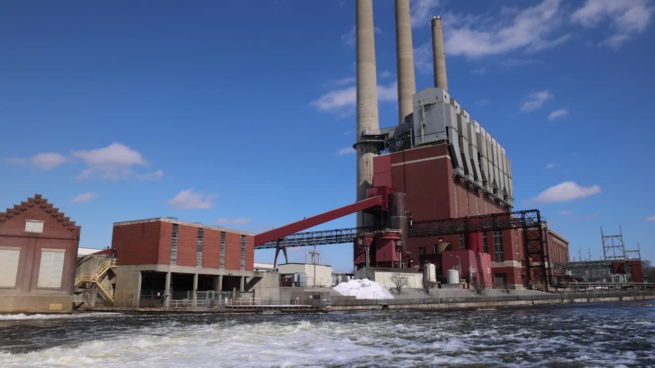 Panning shot from river dam to coal burning power plant with three smokestacks.