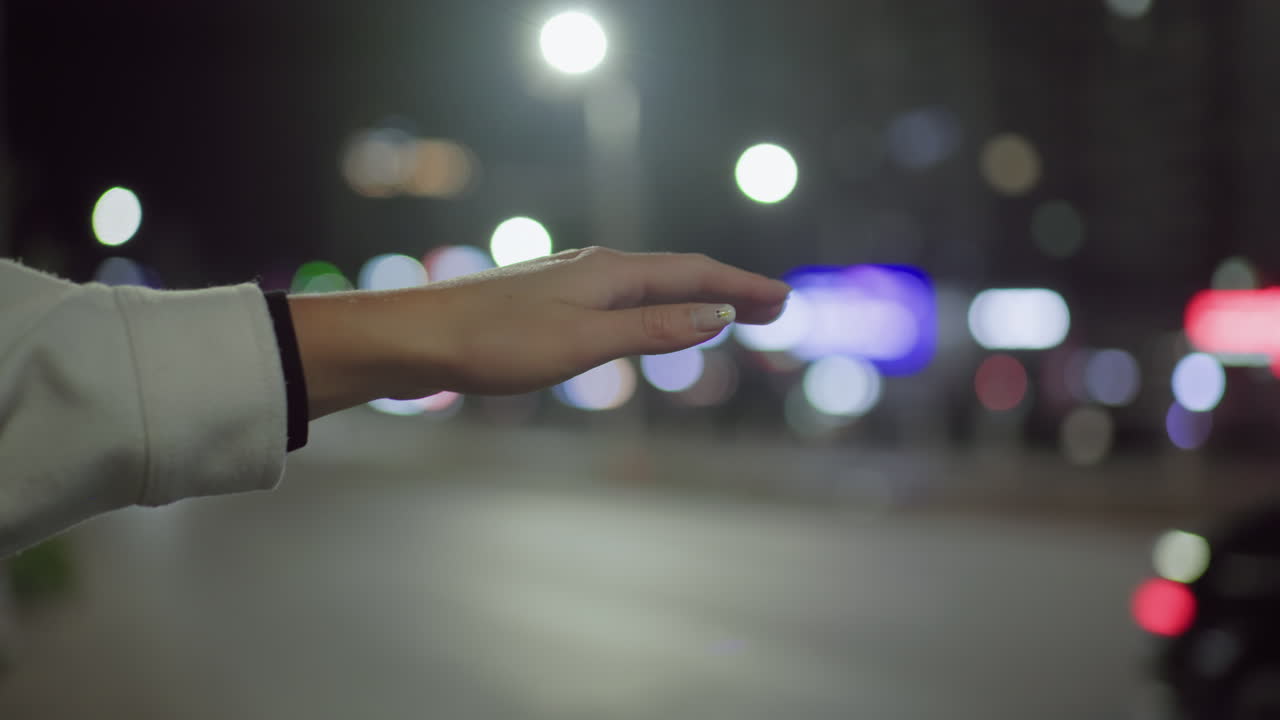 Close-up of female hand in white long sleeve with polished nail extended to stop taxi at night, illuminated by streetlights as car headlights and colorful bokeh light blur fill urban background