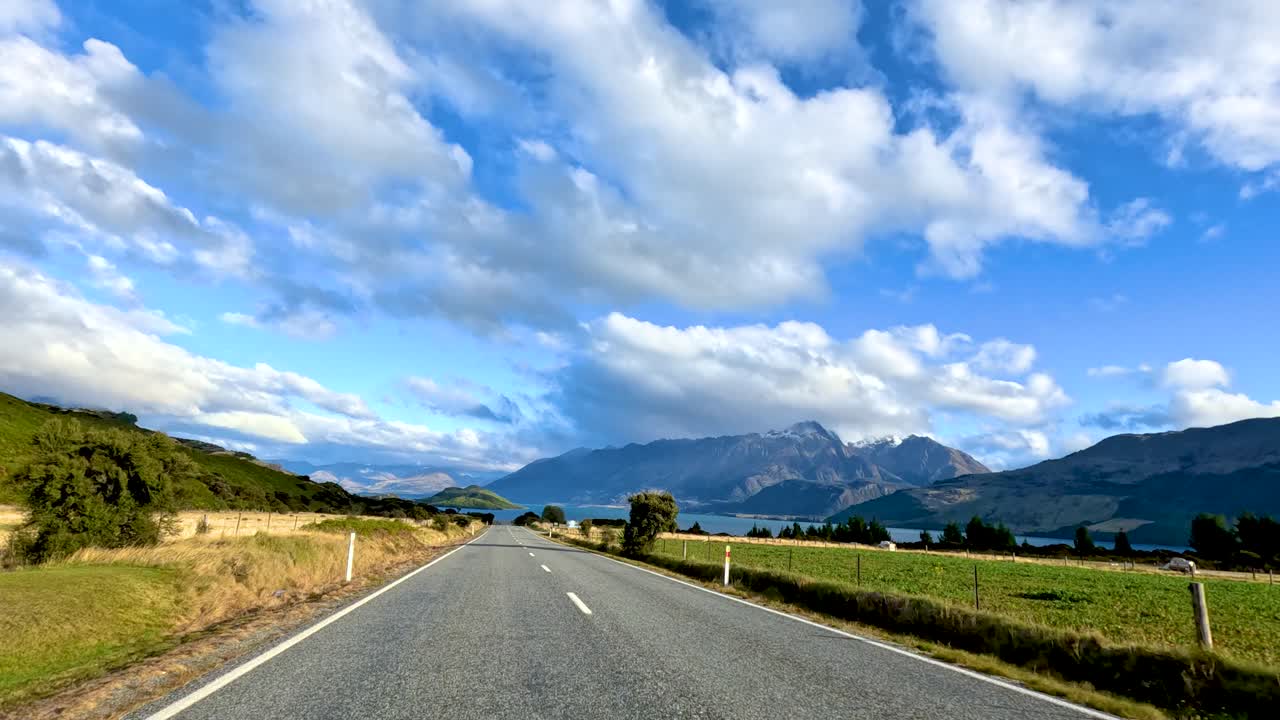 Car drives scenic country road toward mountains and lake under bright daylight, wide angle perspective