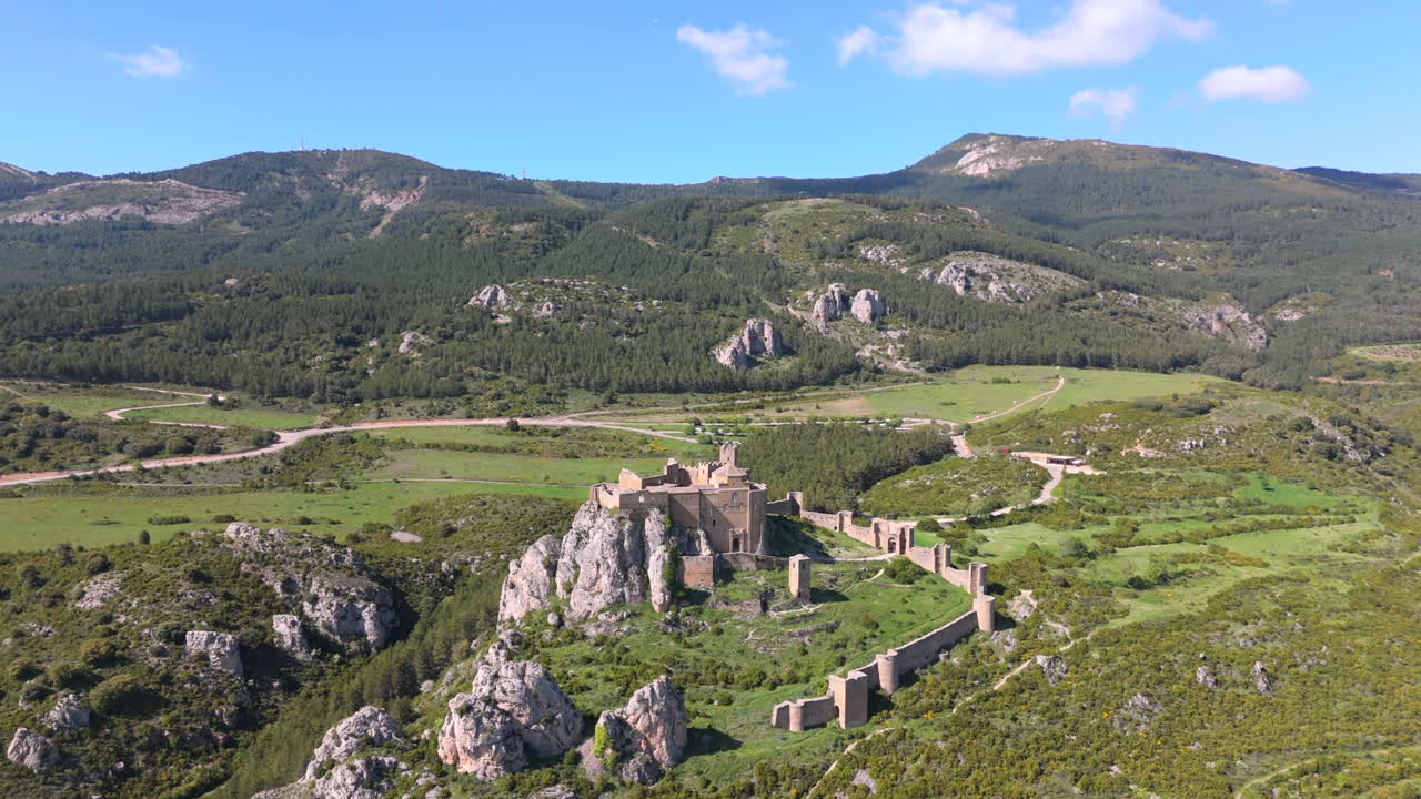 Aerial footage approaching a historic medieval castle on a rocky outcrop, surrounded by dense forest, valleys, and high mountain terrain under a bright blue sky