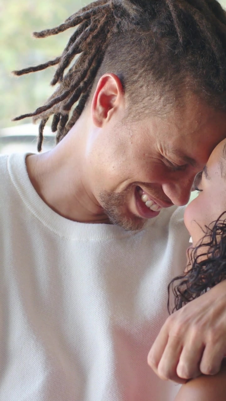 Vertical video: Leaning in man with locs lovingly cradling her near window, white top red top