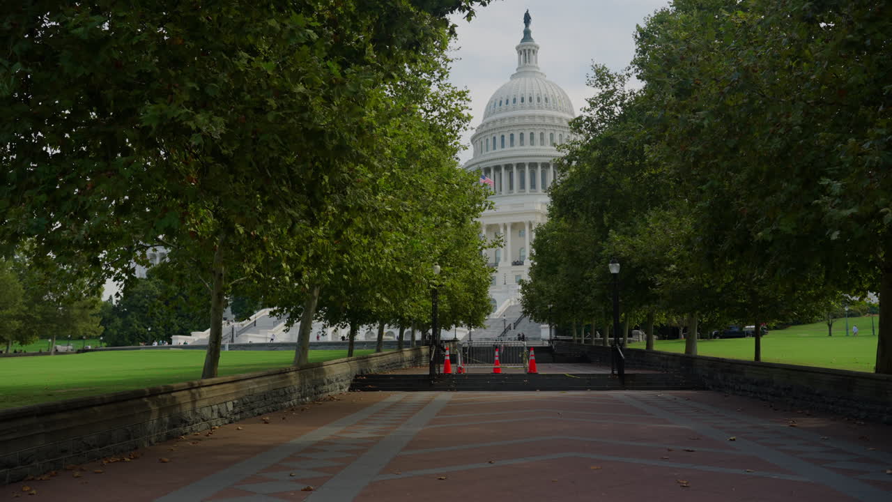 Pathway lined with trees leading to the U.S. Capitol, showing barriers, warning signs, and cones for security measures