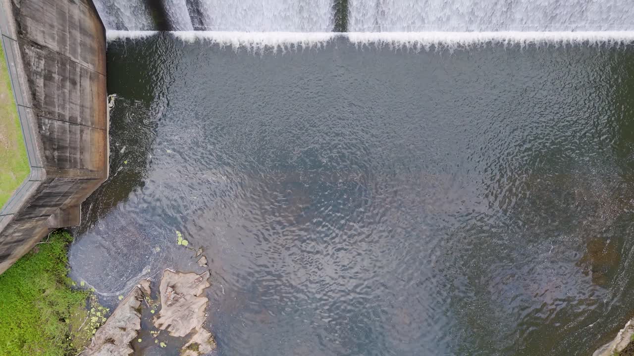 Overhead drone footage captures water cascading over Tallebudgera Dam, surrounded by lush greenery and rocky terrain
