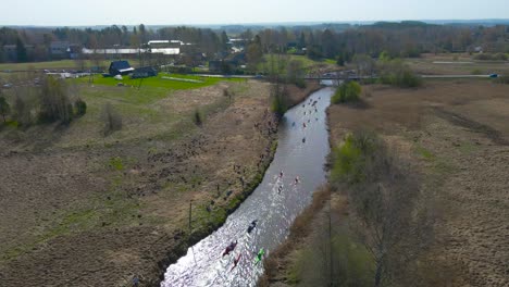 aerial back view of large group of people paddling along river during võhandu marathon. drone rotates above scenic river with spring sunlight while kayaks, canoes on long-distance waterway competition