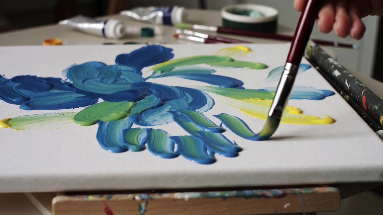 Intingendo il pennello, la mano destra dell'artista dipinge un piccolo motivo floreale su una tela da tavolo, con tubetti di pittura.