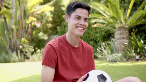 Hombre-Birracial-Feliz-Sentado-Con-Fútbol-Después-De-Entrenar-En-Un-Jardín-Soleado,-Espacio-Para-Copiar,-Cámara-Lenta