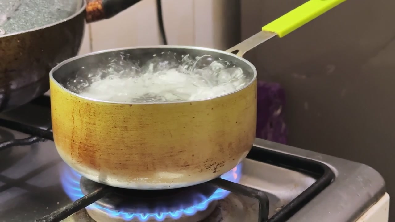 Close-up of a small metal pot boiling water on a gas stove burner. Bubbles and steam rise from the pot as a blue flame heats the bottom in a home kitchen setting.