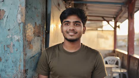 young man with dark hair leaning against weathered blue wall, wearing olive t shirt, smiling softly with closed eyes, embodying peaceful contentment and serene inner harmony