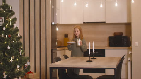 la cámara enfoca a una chica que coloca un vaso de leche y un plato lleno de galletas en una mesa vacía con dos velas en una habitación decorada con un árbol de navidad, luego sale de la habitación