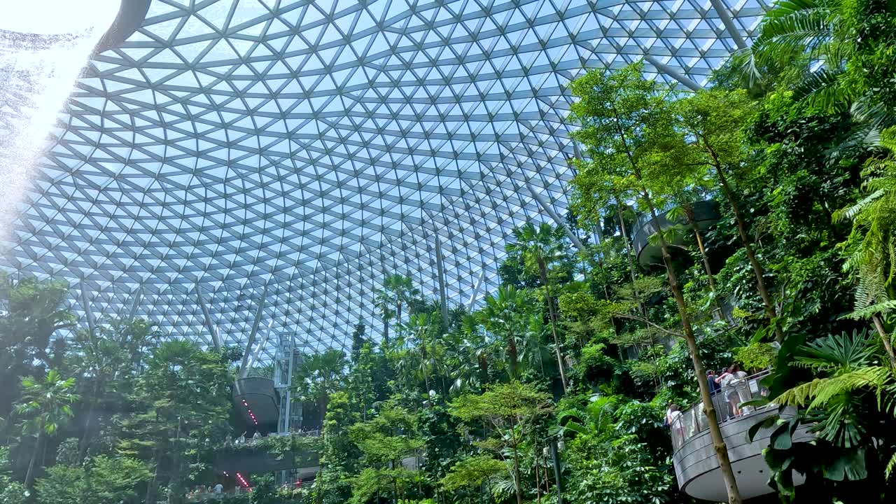 Tall indoor waterfall pours from glass dome into vibrant green garden, bright daylight, wide angle