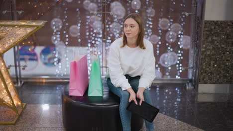 thoughtful young woman seated in a mall holding a tablet, surrounded by shopping bags and festive decorations, gazing away with twinkling lights and a large decorative star