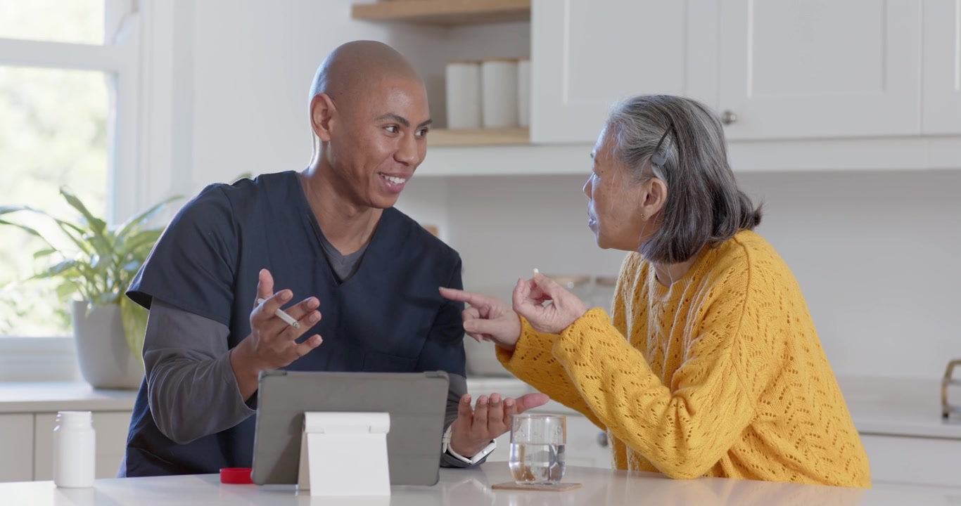 Nurse in scrubs pointing stylus explaining dose on tablet in kitchen as senior woman holding pill