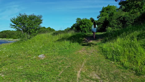 Brunette-woman-wearing-a-white-skirt-walking-along-a-path-between-trees-and-a-lake-on-the-background