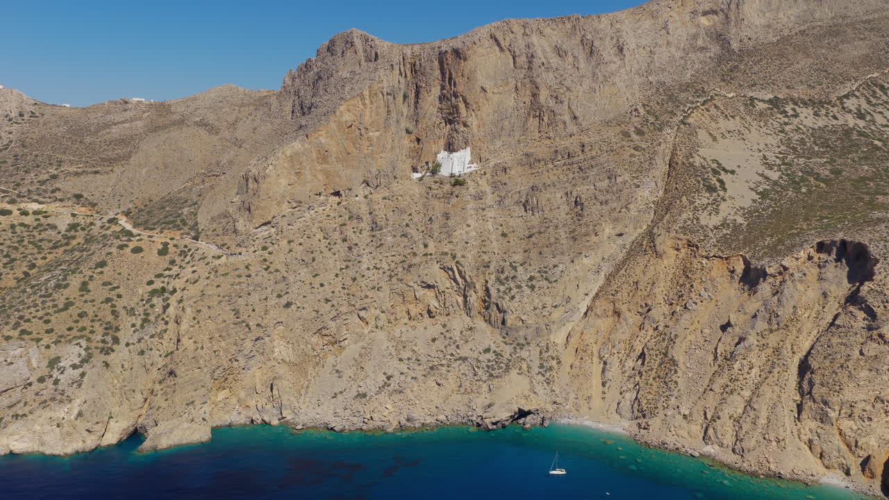 White monastery perched on cliffs above turquoise sea, Amorgos Island, Greece, panoramic aerial orbit establish