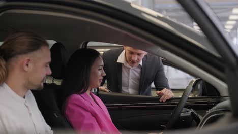 young car salesman showing to young couple new automobile at dealership salon.