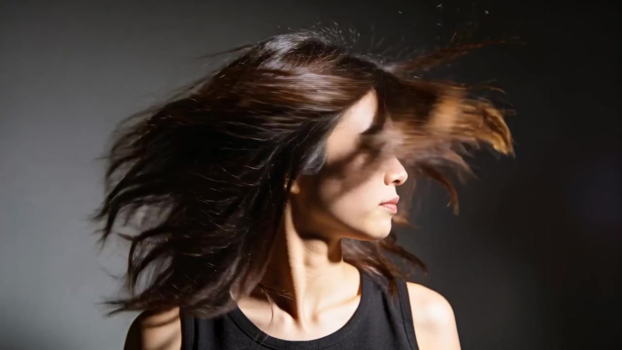 Donna che gira rapidamente la testa, sbatte i lunghi capelli a strati sul viso in studio, indossando una canotta nera.