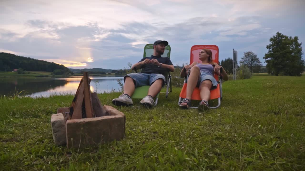 Coppia seduta su sedie da esterno vicino a un falò in riva a un lago calmo al tramonto, che parla e si gode la tranquilla atmosfera del campeggio nell'ambiente naturale di Savci, Slovenia.