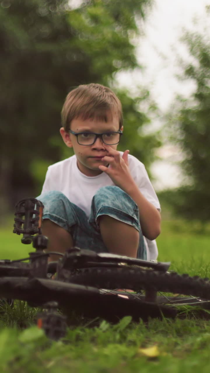 un niño se agacha al lado de su bicicleta, quitó algo de su cara en un campo de hierba con vegetación borrosa en el fondo