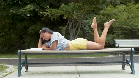Beautiful Latina Woman Reading Book While Fixing Hair And Laying On Stomach On Park Bench
