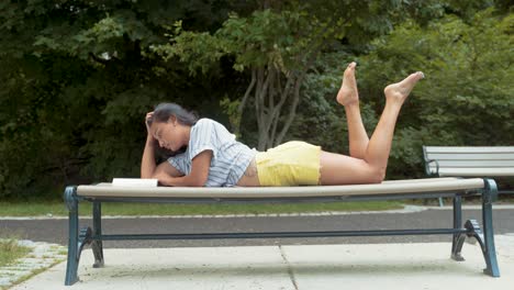 beautiful latina woman reading book while fixing hair and laying on stomach on park bench